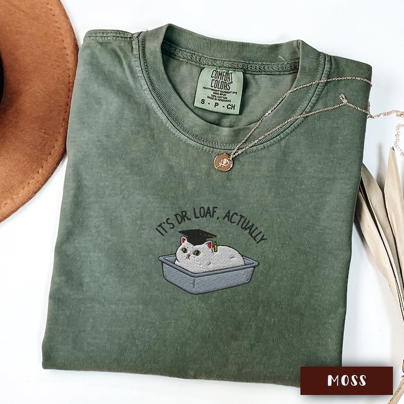 May include: A moss green t-shirt featuring the embroidered text "IT'S DR. LOAF, ACTUALLY" and a cat wearing a graduation cap. The shirt has a crew neck and short sleeves. A gold necklace is draped across the shirt.
