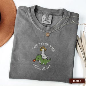 May include: A gray Comfort Colors t-shirt with the embroidered phrase "Off to be the Cool Aunt." The design features a goose wearing a cowboy hat riding a green dinosaur. A silver necklace with a pendant is draped across the shirt.