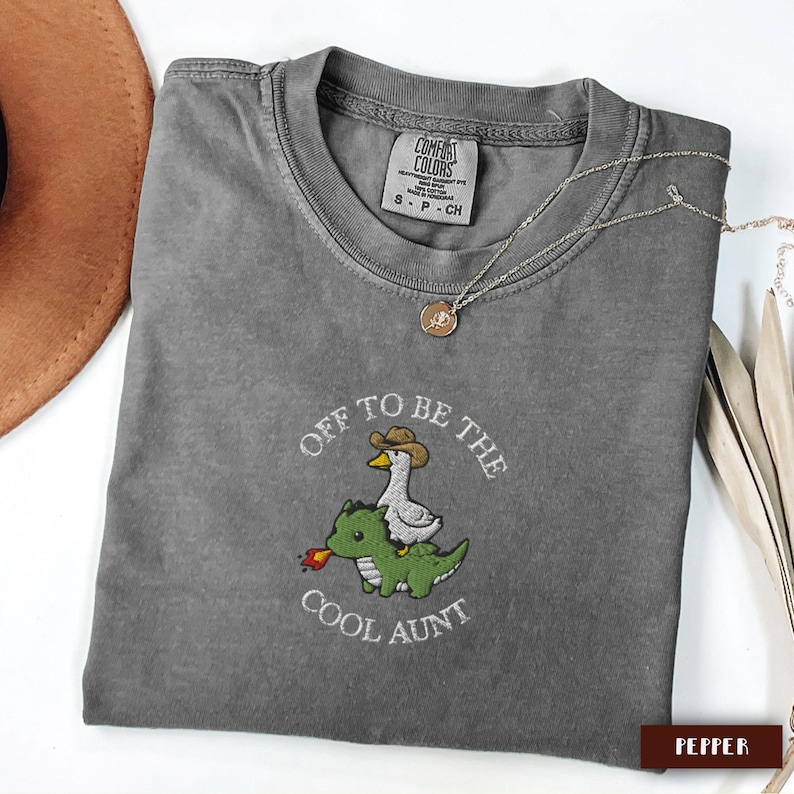 May include: A gray Comfort Colors t-shirt with the embroidered phrase "Off to be the Cool Aunt." The design features a goose wearing a cowboy hat riding a green dinosaur. A silver necklace with a pendant is draped across the shirt.