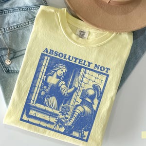以下が含まれることがあります： 中世のシーンの青いグラフィックと「ABSOLUTELY NOT」という言葉がプリントされたバター色のTシャツ。Tシャツは青いジーンズとベージュの帽子の上に折りたたまれています。金のネックレスが見えます。