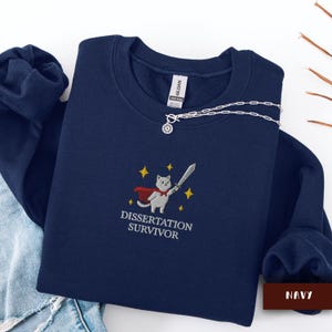 May include: Navy blue sweatshirt with an embroidered design featuring a cat in a cape holding a sword, with the text "DISSERTATION SURVIVOR". A silver necklace is draped across the sweatshirt.