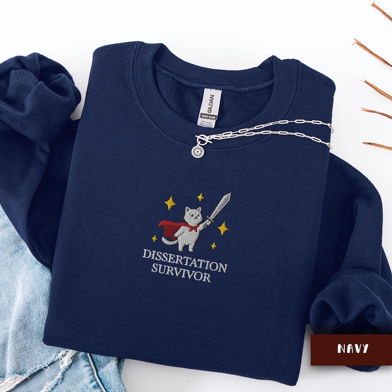 May include: Navy blue sweatshirt with an embroidered design featuring a cat in a cape holding a sword, with the text "DISSERTATION SURVIVOR". A silver necklace is draped across the sweatshirt.