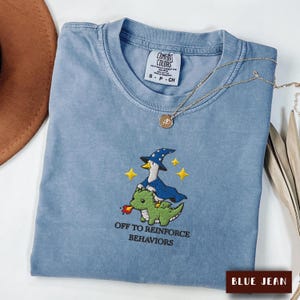 May include: A blue jean coloured t-shirt featuring an embroidered graphic of a goose wearing a blue wizard hat and cape riding a green dragon. The text "OFF TO REINFORCE BEHAVIORS" is embroidered below the graphic. A gold necklace rests on the collar.