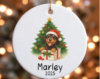 May include: A white ceramic Christmas ornament featuring a dachshund puppy wearing a Santa hat, emerging from a gift box in front of a Christmas tree. The ornament has the name "Marley" and the year "2025" printed on it.