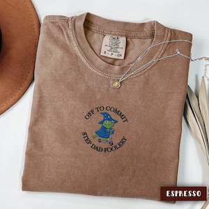 May include: Espresso-colored t-shirt with the phrase "OFF TO COMMIT STEP DAD FOOLERY" embroidered on the front. The graphic features a frog wearing a wizard hat and cape on a scooter. A silver necklace with a pendant is draped across the shirt.
