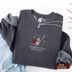 May include: Dark heather gray sweatshirt with an embroidered design of a cat in a cape holding a sword, with the text "Dissertation Survivor". A silver necklace with a pendant is draped across the sweatshirt.