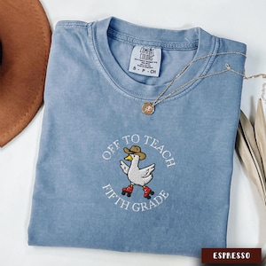 May include: A light blue Comfort Colors t-shirt with the embroidered phrase "Off to Teach Fifth Grade" and a cartoon duck wearing a cowboy hat and roller skates. A gold necklace and a brown hat are also visible.