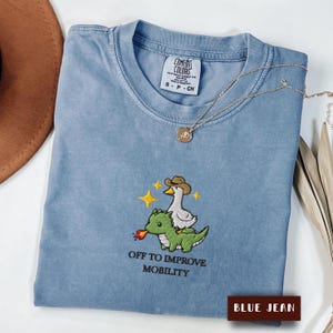 May include: A blue jean colored t-shirt featuring an embroidered graphic of a white duck wearing a brown cowboy hat riding a green dragon that is breathing fire. Above the dragon are yellow stars. The text reads OFF TO IMPROVE MOBILITY.