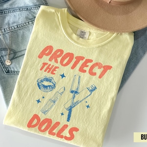 Peut inclure: Un t-shirt jaune clair avec l'inscription "PROTECT THE DOLLS" en rouge, ainsi que des graphiques de rouge à lèvres, de mascara et d'étoiles bleus. Le t-shirt est posé sur un jean bleu et un chapeau beige.