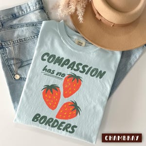 May include: A light blue t-shirt with the words "COMPASSION has no BORDERS" in green, and three red strawberries. The shirt is folded on top of blue jeans, with a tan hat and a gold necklace.