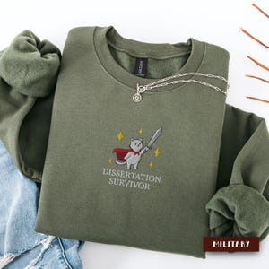 Könnte beinhalten: Ein olivgrünes Sweatshirt mit dem gestickten Bild einer Katze mit Umhang und Schwert, mit dem Text "DISSERTATION SURVIVOR". Eine silberne Halskette mit Sonnenanhänger liegt auf dem Sweatshirt.