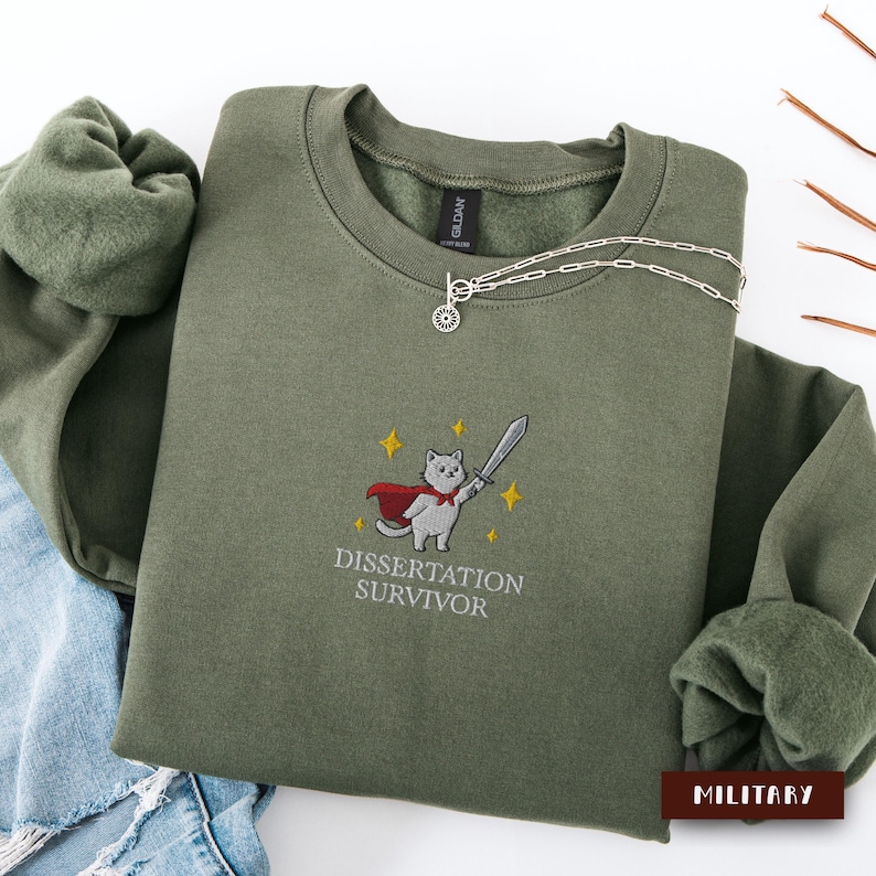 May include: An olive green sweatshirt with the embroidered image of a cat wearing a cape and holding a sword, with the text "DISSERTATION SURVIVOR". A silver necklace with a sun pendant is draped on the sweatshirt.
