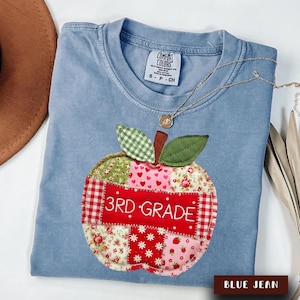 May include: A blue jean colored t-shirt with a Comfort Colors label and a size S tag. The shirt features a large, quilted-style apple graphic made of various red, white, and green patterned fabric patches with the text 3RD GRADE in white.