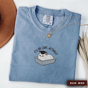 May include: Blue jean-colored Comfort Colors t-shirt with the embroidered phrase "IT'S DR. LOAF, ACTUALLY" and a cat wearing a graduation cap. A gold necklace and a brown hat are also visible.