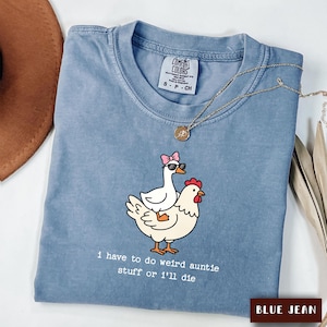 Puede incluir: Una camiseta azul con un gráfico de un ganso blanco con un lazo rosa y gafas de sol montado sobre una gallina blanca. El texto debajo dice, I have to do weird auntie stuff or i'll die. La camiseta tiene una etiqueta de Comfort Colors.