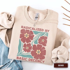 Op de afbeelding: Een zandkleurige sweatshirt met de tekst "RADICALIZED BY BASIC DECENCY" en een bloemenontwerp in rood en blauwgroen. Een zilveren ketting met een kleine hanger ligt op de sweatshirt. De sweatshirt is opgevouwen met een spijkerbroek en een beige stof.