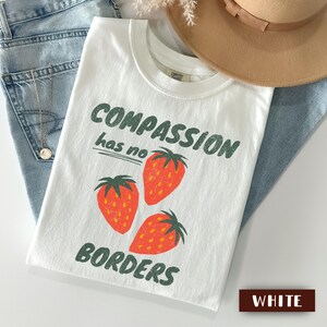 May include: White t-shirt with the text "COMPASSION has no BORDERS" in green, featuring three red strawberries. The shirt is displayed on a light blue denim surface, with a tan hat and a pair of jeans.