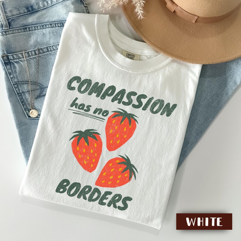 May include: White t-shirt with the text "COMPASSION has no BORDERS" in green, featuring three red strawberries. The shirt is displayed on a light blue denim surface, with a tan hat and a pair of jeans.