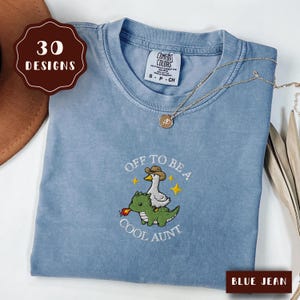 May include: A blue jean coloured t-shirt featuring an embroidered graphic of a white goose wearing a brown cowboy hat riding a green dragon that is breathing fire. The text reads OFF TO BE A COOL AUNT with yellow stars surrounding the design.