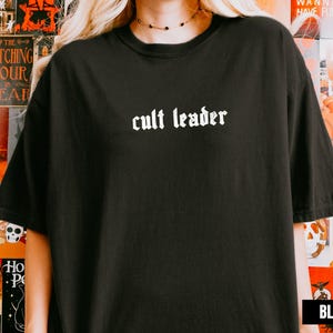 Op de afbeelding: Zwart T-shirt met de tekst "cult leader" in een gotisch lettertype. Het shirt is effen zwart en heeft een ronde hals. De tekst is wit. Het shirt heeft een casual stijl.