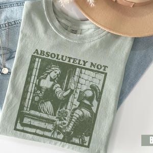 以下が含まれることがあります： 「ABSOLUTELY NOT」の文字と女性と騎士のグラフィックがプリントされた、薄い緑色のTシャツ。Tシャツは、青いジーンズ、ネックレス、ベージュの帽子の上に置かれています。