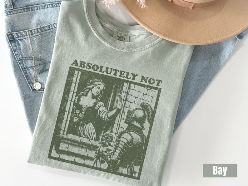 以下が含まれることがあります： 「ABSOLUTELY NOT」の文字と女性と騎士のグラフィックがプリントされた、薄い緑色のTシャツ。Tシャツは、青いジーンズ、ネックレス、ベージュの帽子の上に置かれています。