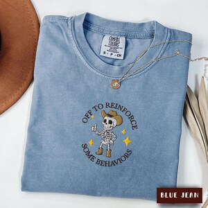 May include: A blue jean coloured t-shirt with a crew neckline and a Comfort Colors label. The shirt features an embroidered graphic of a skeleton wearing a cowboy hat and boots, giving a thumbs up, with the text OFF TO REINFORCE SOME BEHAVIORS.
