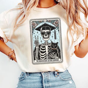 May include: A white t-shirt featuring a graphic of a skeleton wearing a graduation cap and sunglasses, holding a black cat. The graphic is in a tarot card style with the text "The Dissertation Defense Survivor".
