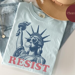 May include: Light blue t-shirt featuring a graphic of the Statue of Liberty wearing heart-shaped sunglasses and blowing a bubble. The word "RESIST" is printed in red. The shirt is folded on top of blue jeans and a tan hat.
