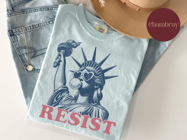 May include: Light blue t-shirt featuring a graphic of the Statue of Liberty wearing heart-shaped sunglasses and blowing a bubble. The word "RESIST" is printed in red. The shirt is folded on top of blue jeans and a tan hat.