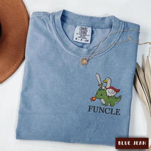 May include: A blue short-sleeved t-shirt with a Comfort Colors label at the neck. The shirt features an embroidered graphic of a white duck wearing a red cape and holding a sword while riding a green dragon breathing fire, with the text FUNCLE below.