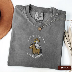 May include: A gray Comfort Colors t-shirt with the embroidered phrase "Off to be the Cool Aunt." The design features a horse and a duck wearing a cowboy hat. A silver necklace with a pendant is draped across the shirt.