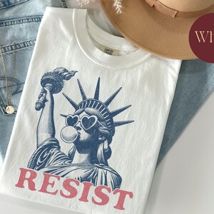 May include: White t-shirt featuring a blue graphic of the Statue of Liberty wearing heart-shaped sunglasses and blowing a bubble. The word "RESIST" is printed in red below the image. The shirt is folded on top of blue jeans.