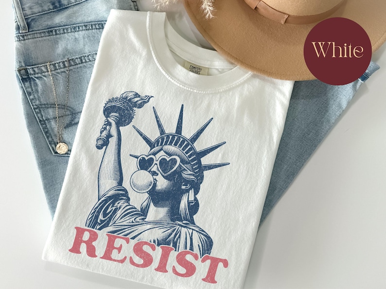 May include: White t-shirt featuring a blue graphic of the Statue of Liberty wearing heart-shaped sunglasses and blowing a bubble. The word "RESIST" is printed in red below the image. The shirt is folded on top of blue jeans.