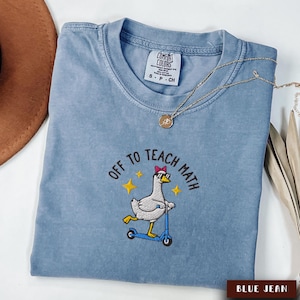 May include: Blue jean Comfort Colors t-shirt with an embroidered design. The design features a goose wearing sunglasses and a bow riding a scooter, with the text "OFF TO TEACH MATH". A gold necklace is draped across the shirt.
