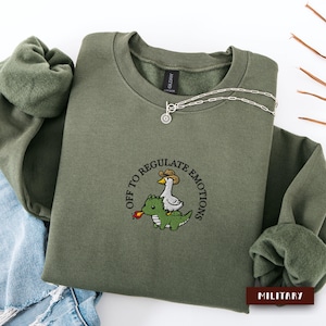 Può includere: Una felpa girocollo verde oliva con un ricamo che raffigura un'oca bianca con un cappello da cowboy marrone a cavallo di un drago verde che sputa una piccola fiamma arancione. Il testo "OFF TO REGULATE EMOTIONS" circonda il disegno.