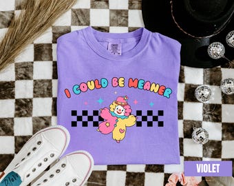 May include: A violet t-shirt with the phrase "I COULD BE MEANER" and a cartoon clown graphic. The clown is pink and yellow with a checkered border. White sneakers are at the bottom of the image.