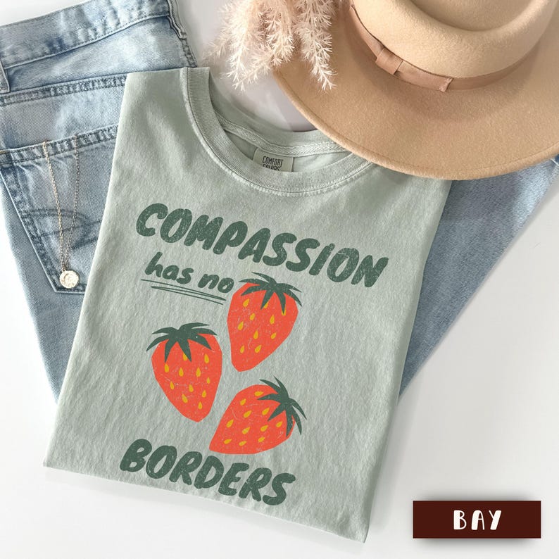 May include: A light green t-shirt with the words "COMPASSION has no BORDERS" and three orange strawberries. The shirt is paired with blue jeans and a tan hat. The shirt is a Comfort Colors brand.