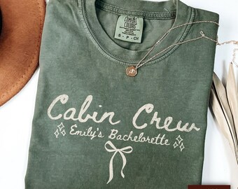 May include: A moss green t-shirt with the words "Cabin Crew" and "Emily's Bachelorette" printed in white script. A gold necklace with a pendant rests on the shirt. A brown hat is visible in the upper left corner.