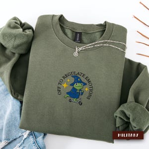 Puede incluir: Una sudadera de cuello redondo color verde oliva con un bordado de una rana verde con sombrero y túnica de mago azul montada en un patinete plateado. El texto "OFF TO REGULATE EMOTIONS" aparece arqueado sobre la rana con estrellas amarillas.