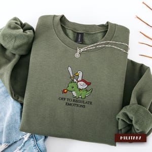 Peut inclure: Un sweat-shirt à col rond vert olive avec une broderie représentant une oie blanche portant une cape rouge et tenant une épée tout en chevauchant un dragon vert. Sous le motif, le texte OFF TO REGULATE EMOTIONS est écrit en lettres noires.