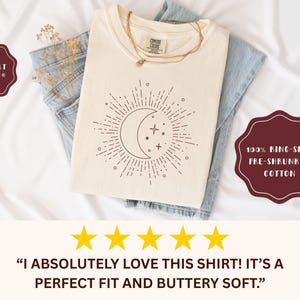 May include: Cream-colored t-shirt with a celestial design featuring a crescent moon, stars, and sunburst. The shirt is folded over blue jeans. The text on the shirt reads "COMFORT COLORS" and "100% RING-SPUN PRE-SHRUNKEN COTTON".