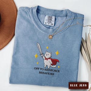 May include: A blue jean coloured t-shirt featuring an embroidered graphic of a white cat wearing a red cape and holding a sword, surrounded by yellow stars. The text below the cat reads OFF TO REINFORCE BEHAVIORS. The shirt has a crew neckline.
