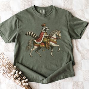 May include: A green t-shirt with a graphic of a raccoon wearing a crown and riding a white horse. The raccoon is wearing a red and gold robe.