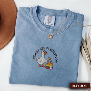 May include: A blue jean Comfort Colors t-shirt with the embroidered phrase "Dissertation Survivor" around a cartoon duck wearing sunglasses and holding a match to a stack of books. A gold necklace is draped across the shirt.