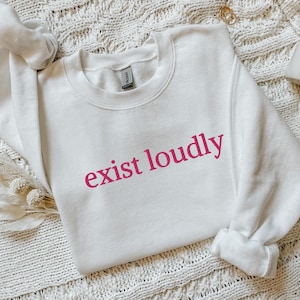 May include: White crewneck sweatshirt with the phrase "exist loudly" printed in pink.