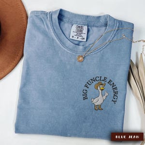 May include: Blue jean-colored Comfort Colors t-shirt with the embroidered phrase "BIG FUNCLE ENERGY" surrounding a cartoon goose wearing a cowboy hat and boots. A gold necklace with a pendant is draped across the shirt.