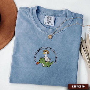 May include: A light blue Comfort Colors t-shirt with an embroidered design featuring a goose wearing a cowboy hat riding a green dinosaur. The text "OFF TO REGULATE EMOTIONS" is embroidered around the design. A gold necklace is draped across the shirt.