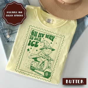 May include: Butter-colored t-shirt with a green graphic of a frog riding a scooter, wearing a wizard hat and cape. The text "ON MY WAY TO HEX ICE" is printed above the frog. The shirt is folded and placed on top of blue jeans and a tan hat.