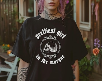 May include: Black t-shirt with the text "Prettiest Girl in the Morgue" in white gothic lettering. The design includes a detailed skull illustration within a circular frame. The shirt is a casual, oversized fit.